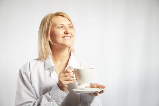 Portrait of pretty blonde smiling woman posing with cup of tea or coffee on white background. Happy girl model in white shirt in studio. The concept of pleasant morning at home or work. Copy space