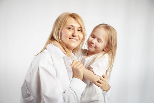 Portrait of blonde mother and daughter who having communicate and play on a white background. Mom and little girl models pose in the studio. The concept of love, friendship, caring in the family