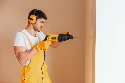 Handyman in yellow uniform works indoors by using hammer drill. House renovation conception.