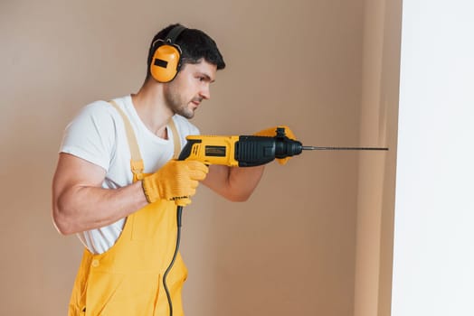 Handyman in yellow uniform works indoors by using hammer drill. House renovation conception.