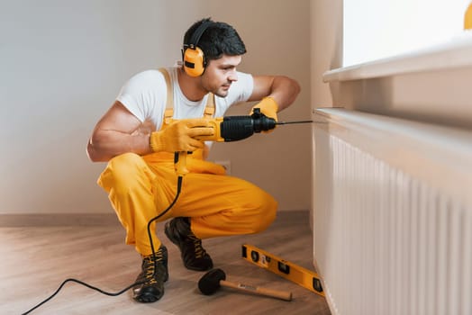 Handyman in yellow uniform works indoors by using hammer drill. House renovation conception.