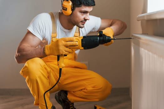 Handyman in yellow uniform works indoors by using hammer drill. House renovation conception.