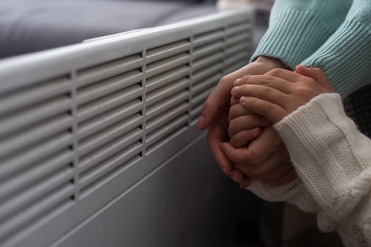 Mother and child warming hands near electric heater at home, closeup