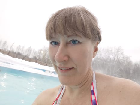 Adult mature woman takes selfie in a pool with warm hot termal mineral water in winter and white snow around. Wellness center and the concept of health care. Travel, recreation, medicine and rest