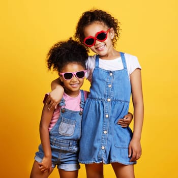 Children, sunglasses and happy sisters in studio hug for love, care and support of family on yellow background. Cute young girl kids portrait together for happiness, carefree and fashion with a smile.