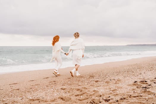 Women sea walk friendship spring. Two girlfriends, redhead and blonde, middle-aged walk along the sandy beach of the sea, dressed in white clothes. Against the backdrop of a cloudy sky and the winter sea. Weekend concept