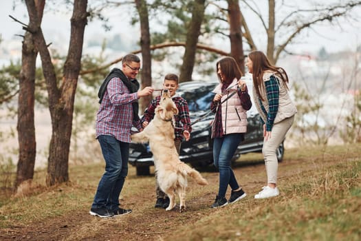 Happy family have fun with their active dog near modern car outdoors in forest.