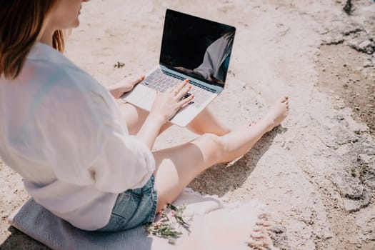 Successful business woman in yellow hat working on laptop by the sea. Pretty lady typing on computer at summer day outdoors. Freelance, travel and holidays concept.