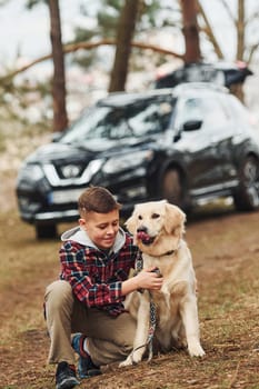 Cheerful boy in casual clothes sitting with her dog in forest against modern black car.