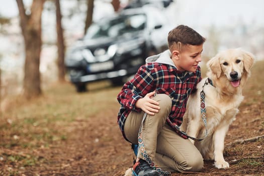 Cheerful boy in casual clothes sitting with her dog in forest against modern black car.