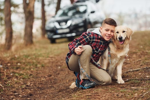 Cheerful boy in casual clothes sitting with her dog in forest against modern black car.