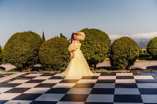 Woman yellow dress chess. A beautiful woman in a long puffy yellow dress poses on a chessboard in the park