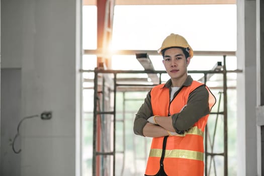 Portrait of asian man architect at building site with folded arms looking at camera. Confident construction manager in formal clothing wearing yellow hardhat. Successful engineer..