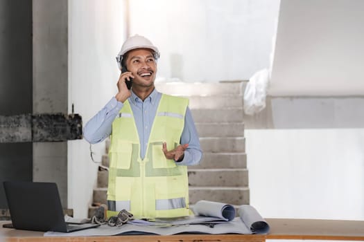 Engineer or Architect with protective safety helmet checking using smartphone holding architectural drawing at construction site. Engineering, Architecture and building project concepts..