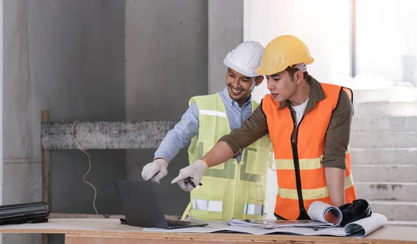 young man construction team engineer in vest and hat working with laptop standing on construction site House building project. Engineer talking with colleagues at work.