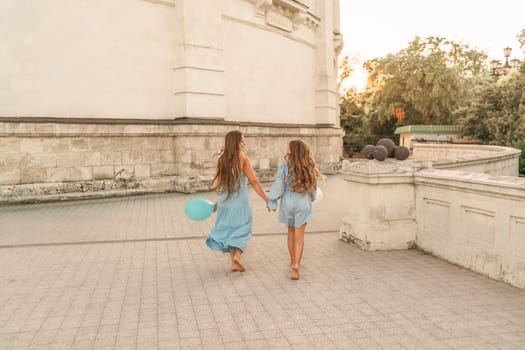 Daughter mother run holding hands. In blue dresses with flowing long hair, they hold balloons in their hands against the backdrop of a sunset and a white building