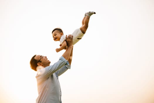In a beautiful moment of family togetherness, a father holds his baby daughter up high, throwing her up into the sunny sky at sunset. The cheerful child smiles with freedom and playful fun