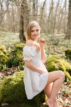 Snowdrops galanthus blonde. A girl in a white dress sits on a meadow with snowdrops in a spring forest.