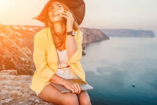 Successful business woman in yellow hat working on laptop by the sea. Pretty lady typing on computer at summer day outdoors. Freelance, travel and holidays concept.