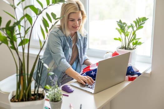 Happy woman employee sitting wrapped in USA flag, shouting for joy in office workplace, celebrating labor day or US Independence day. Indoor studio studio shot isolated on yellow background.