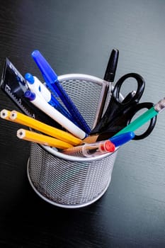 Metal stand with pencils, pens, scissors on a black wooden table close up
