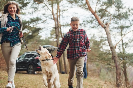 Cheerful girl with brother have walk with their dog outdoors in forest at autumn or spring season near car.