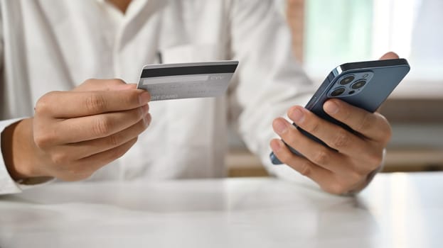 Close up view of man hand holding credit card making transaction using mobile bank application.