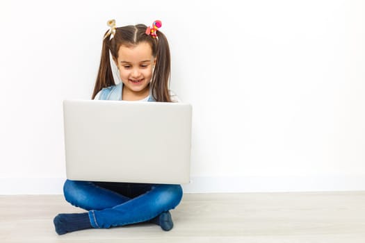 Cute little girl is sitting on floor with her laptop, wearing glasses, isolated over white