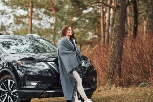 Young cheerful girl standing near modern black car outdoors in the forest.