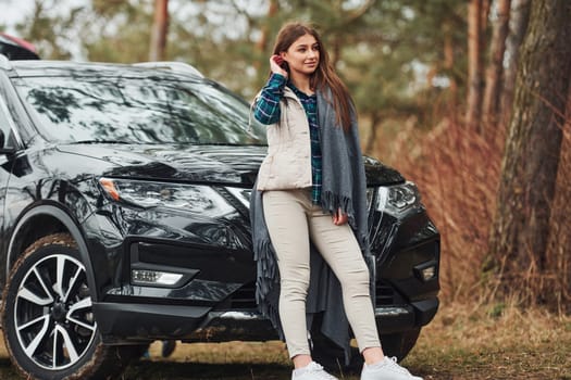 Young cheerful girl standing near modern black car outdoors in the forest.