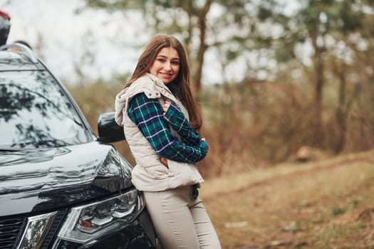 Young cheerful girl standing near modern black car outdoors in the forest.