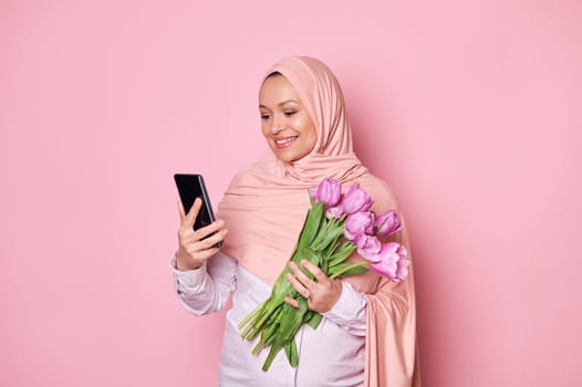 Arab Muslim pregnant woman in pink hijab, holding a bouquet of purple tulips and checking her smartphone, downloading new mobile application, text messaging, smiling on isolated pink color background