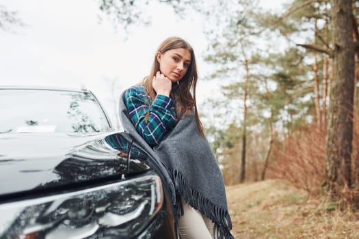 Young cheerful girl standing near modern black car outdoors in the forest.