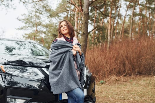 Woman standing near modern black car outdoors in the forest.