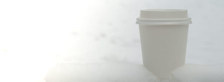 Hot warming drink in Eco paper cup. Mock up Cardboard coffee or tea Cup in the snow in winter day. Copy space Creative trendy zero waste recycle cup white snow-covered field