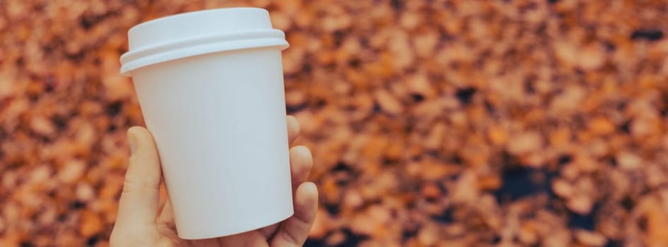 Unrecognizable woman hand holding Eco zero waste white paper cup copy space mockup. Fall leaves and cup of tea coffee to go next to autumn nature. Unite with nature cottagecore