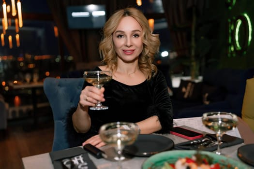 woman in a restaurant with champagne in her hand. She is sitting at a table against the backdrop of a dark restaurant hall with lamps