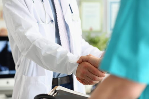 Closeup of two doctors shaking hands against backdrop of medical office. Concept of cooperation in medicine