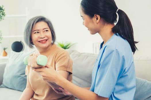 Caregiver helping contented senior woman exercise with dumbbell at home. Rehab for elder, senior medical care.