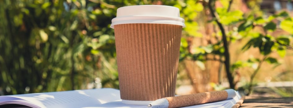 Eco recycling paper cup with coffee or tea on kraft paper with empty paper notebook on wooden bench. Concept of study work outdoors. Take away coffee to go. Copy space for text. Disposable Cardboard coffee outdoors