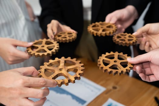 Closeup top view businesspeople hand holding gear and join together over meeting table with financial report papers. Cohesive group of office workers holding cog wheel as synergy harmony concept.