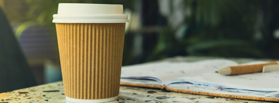 Hot latte coffee in craft recycling paper cup with paper notebook. A take away paper cup on cafe table. Freelance Workspace notebook with coffee for productive work and study. Working comfortably. Empty workspace. Blurred background. Mockup Coffee break. Online job or studying co-working space