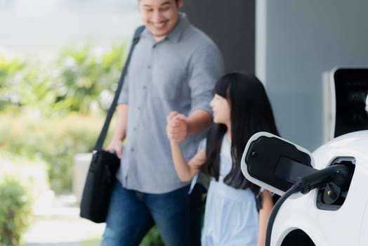 Focus electric car recharging at home charging station with blurred father and daughter walking in background. Progressive green and clean energy vehicle for healthy environment lifestyle concept.