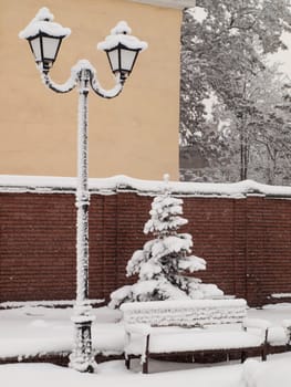 Vintage black street lamppost with two lamps and building, wall, tree and bench covered with snow