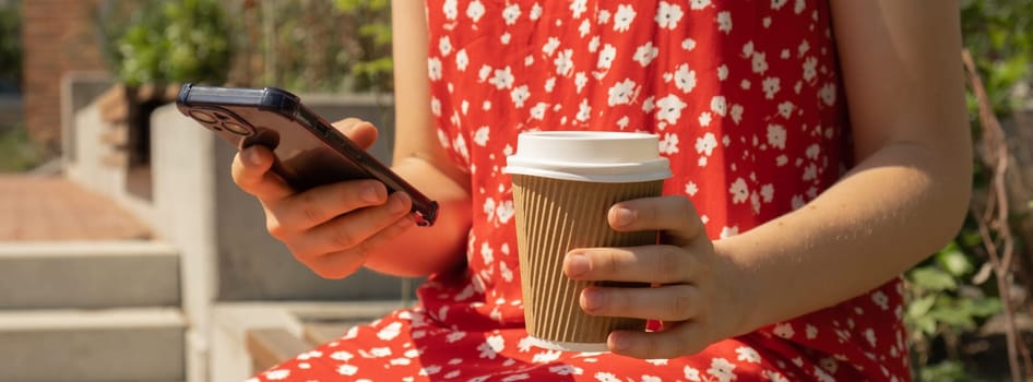 Unrecognizable Young woman scrolling social media on mobile phone on bench in park. Using mobile phone and holding paper recycling cup of coffee while sitting in park on sunny day. Side view of unrecognizable caucasian woman coffee break, chilling outdoors, close up