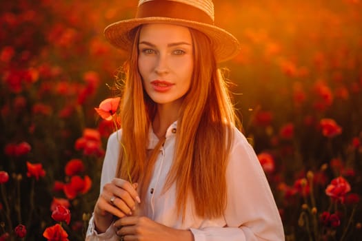 a girl in a white dress and hat stands in a field with poppies at sunset and holds a poppy flower in her hand.