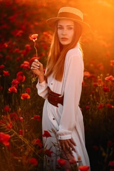 a girl in a white dress and hat stands in a field with poppies at sunset and holds a poppy flower in her hand.