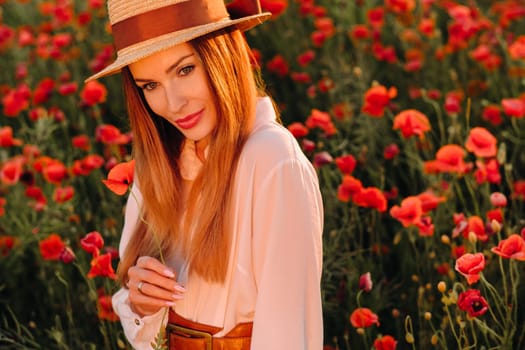 a girl in a white dress and hat stands in a field with poppies at sunset and holds a poppy flower in her hand.