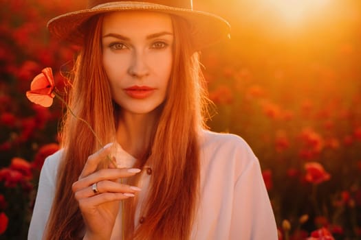a girl in a white dress and hat stands in a field with poppies at sunset and holds a poppy flower in her hand.