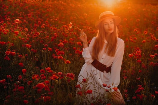 a girl in a white dress and hat stands in a field with poppies at sunset and holds a poppy flower in her hand.